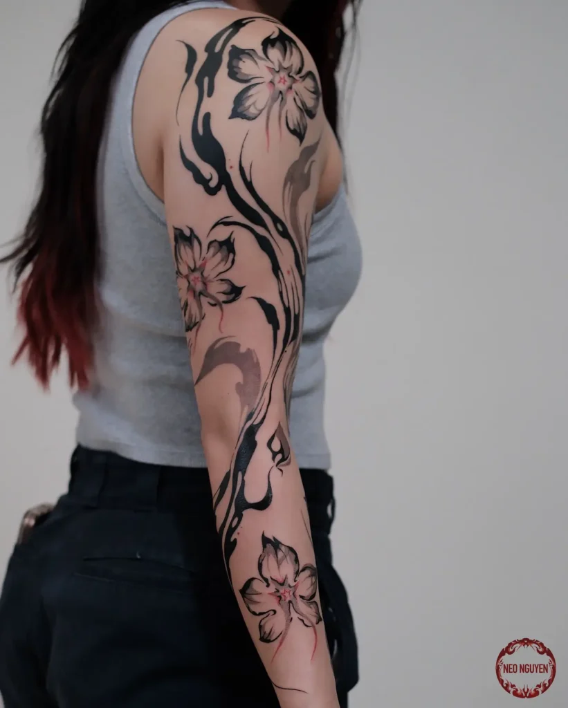 Sleeve tattoo featuring cherry blossoms with black and gray brushstroke patterns and hints of red ink on a woman’s arm.