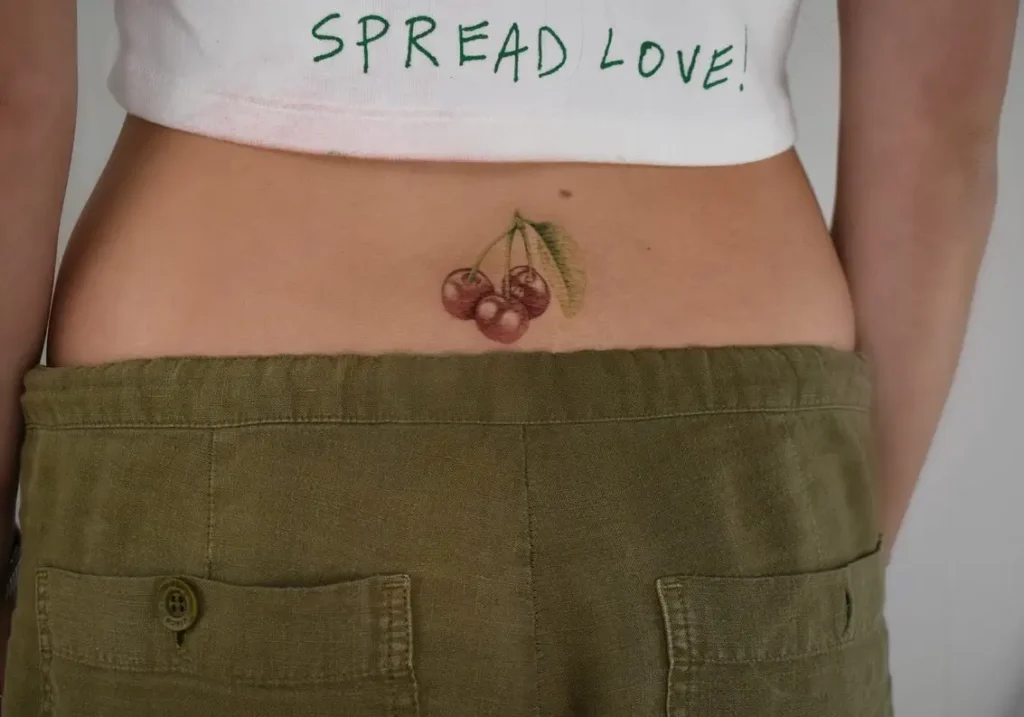  Lower back tattoo with two realistic red cherries and a green leaf, designed with natural shading and color.