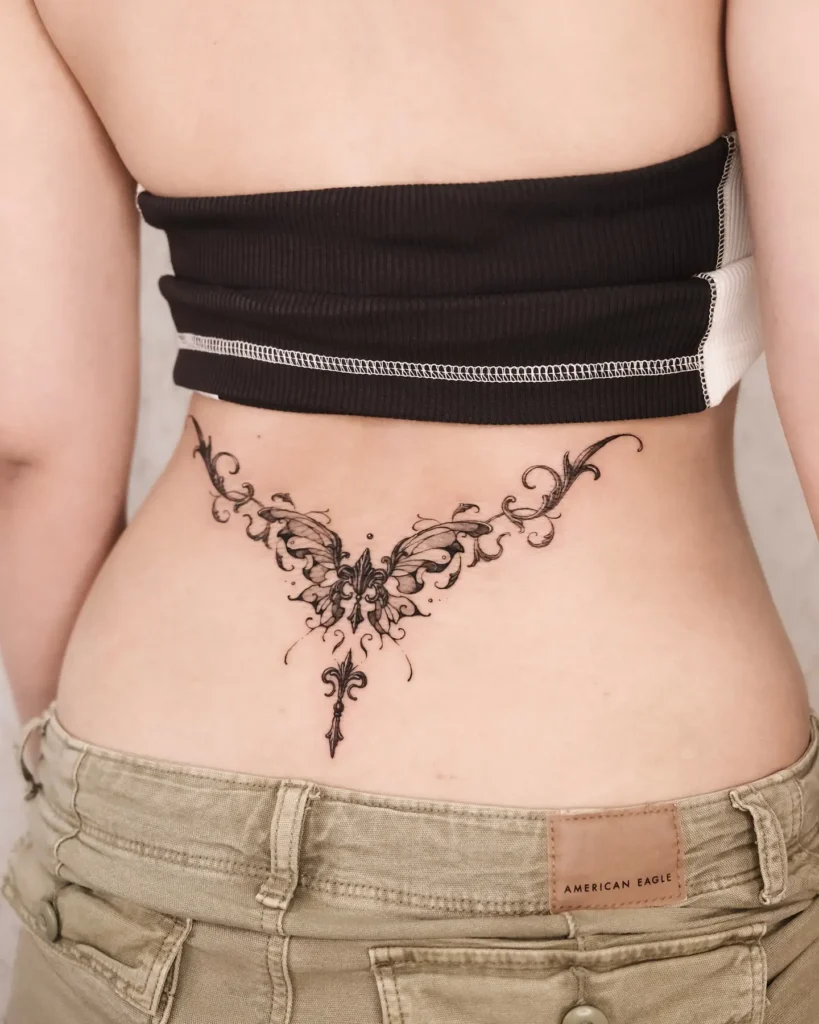  Lower back tattoo of a butterfly with intricate baroque swirls and flourishes, designed in fine black ink.