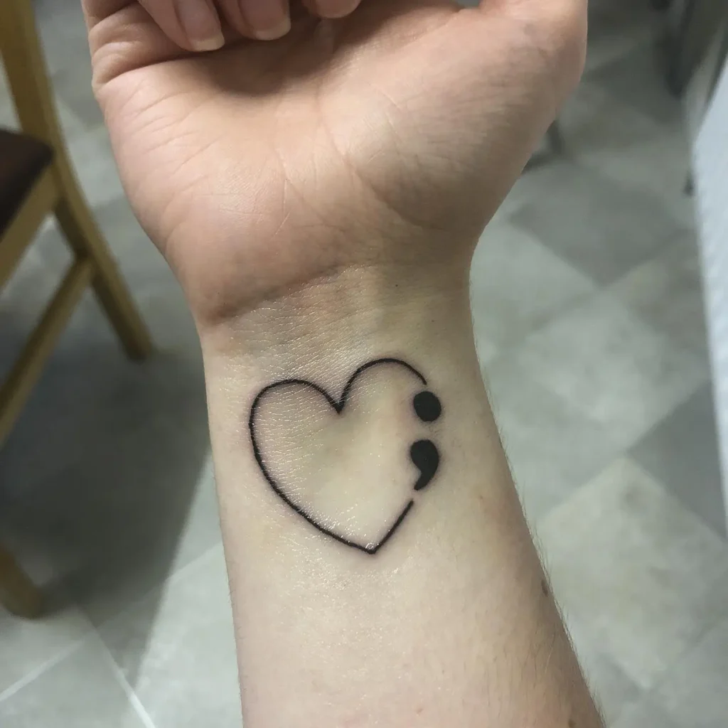A wrist tattoo of a heart outline with a semicolon, representing hope, self-love, and mental health awareness.