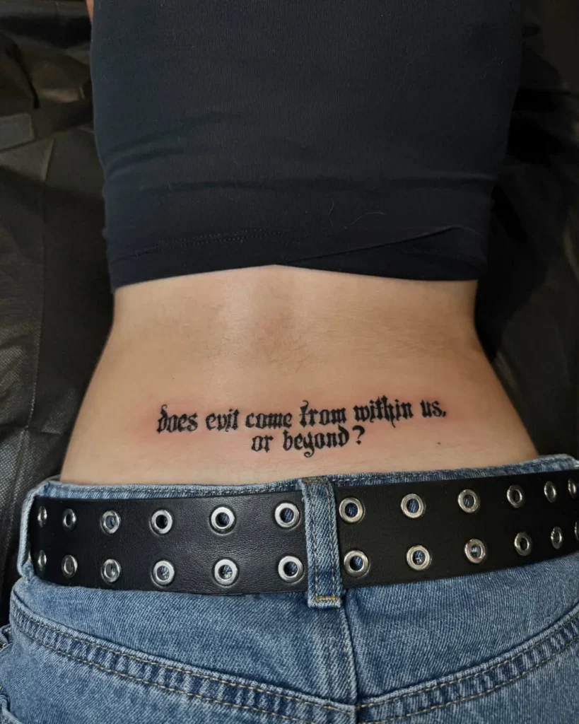 Lower back tattoo of a quote in black gothic script that reads, "does evil come from within us, or beyond?"