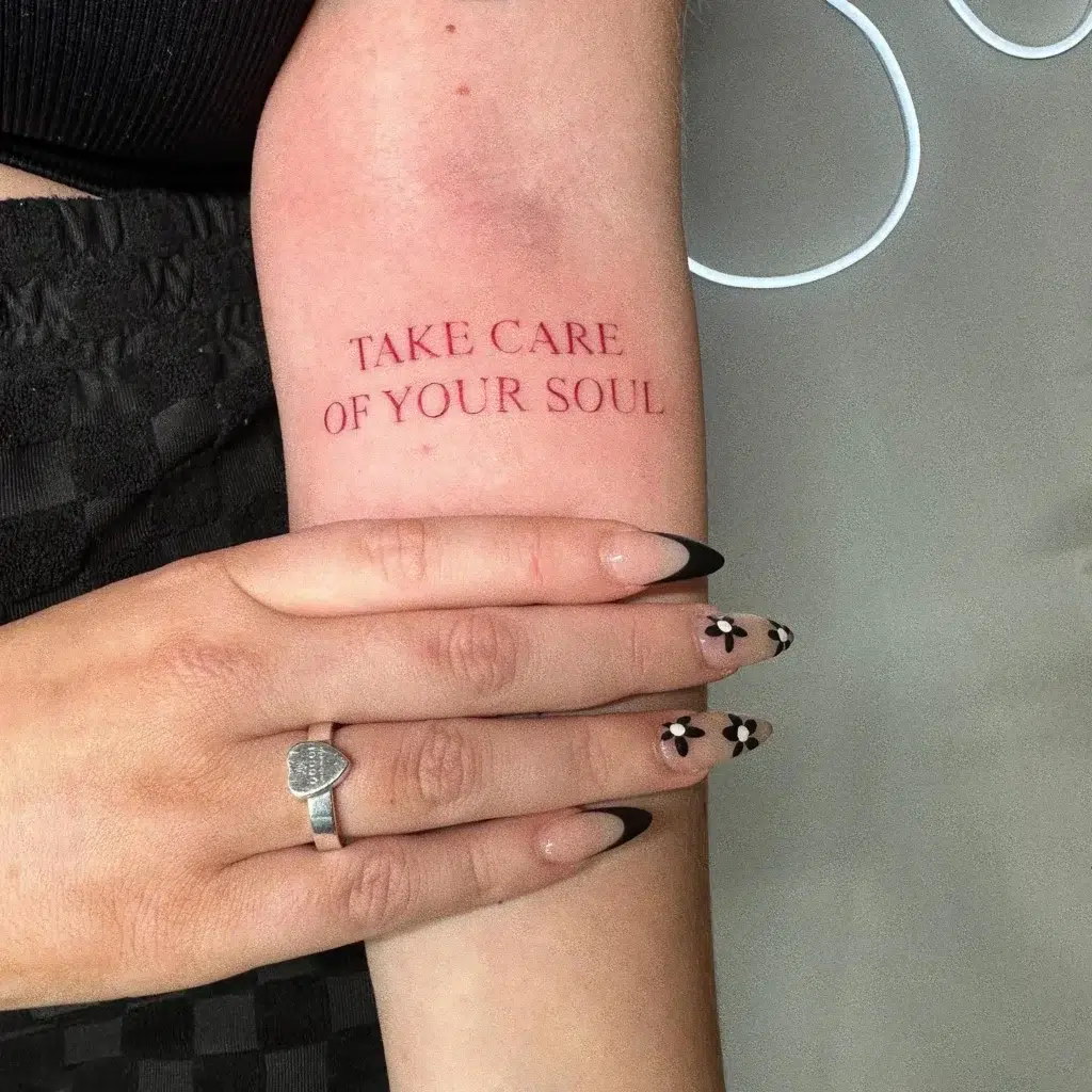 A forearm tattoo in red ink with the phrase “Take care of your soul,” highlighting the importance of self-care and mental well-being.