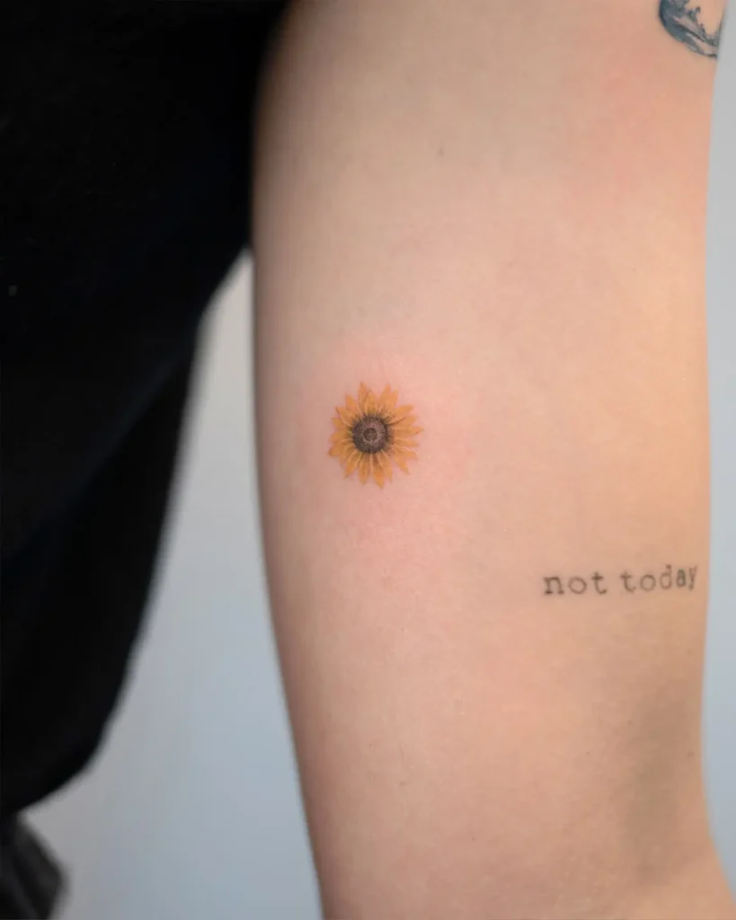 A small sunflower tattoo next to the words “not today” on the upper arm, symbolizing hope, resilience, and self-care.