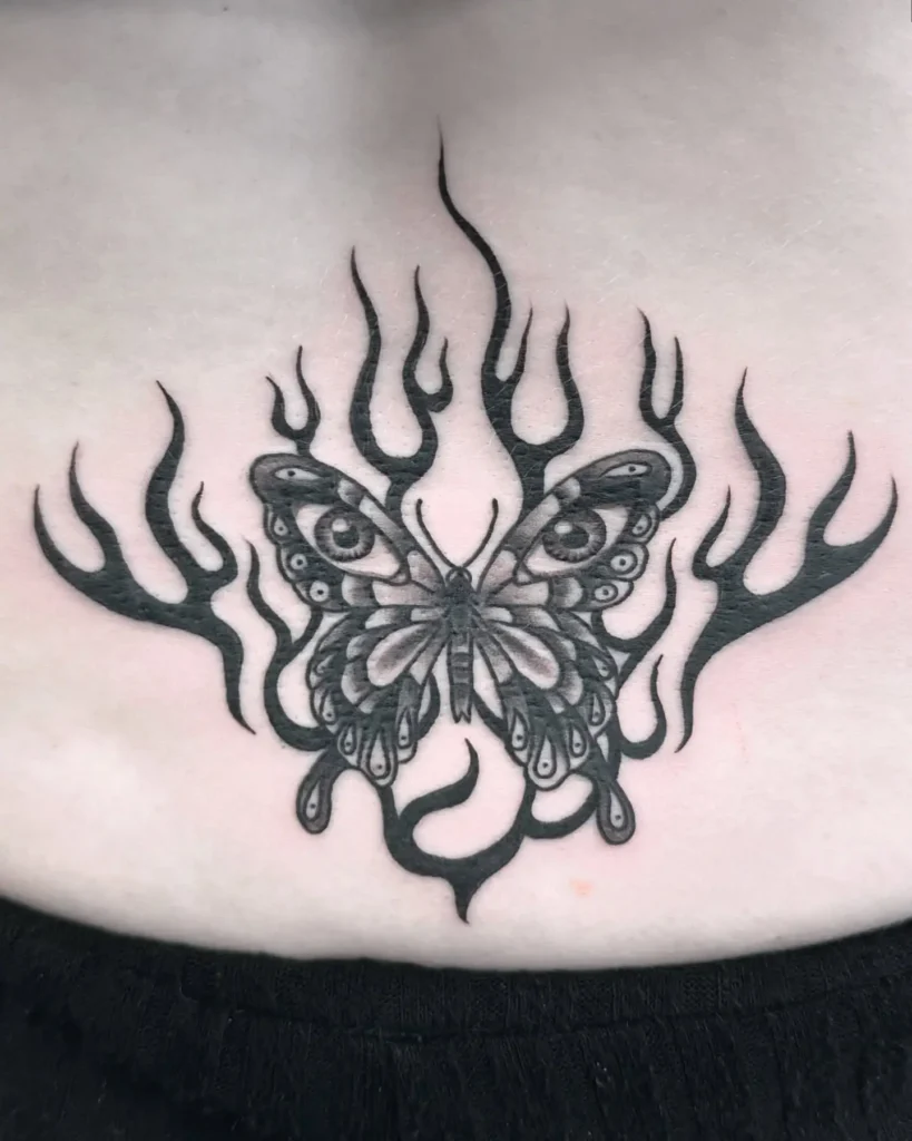 Lower back tattoo of a butterfly with eye patterns on its wings, surrounded by bold black flames.