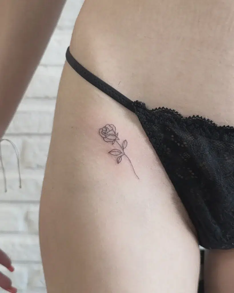 Minimal rose tattoo for women on the upper hip, fine-line floral design placed beneath black lace lingerie—delicate, romantic, and feminine body art.