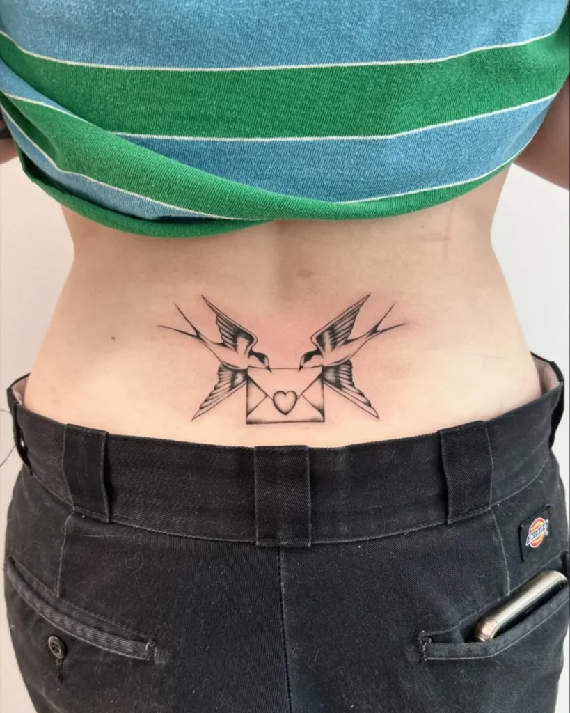Lower back tattoo with two swallows holding a heart-sealed envelope, drawn in fine black and gray lines.