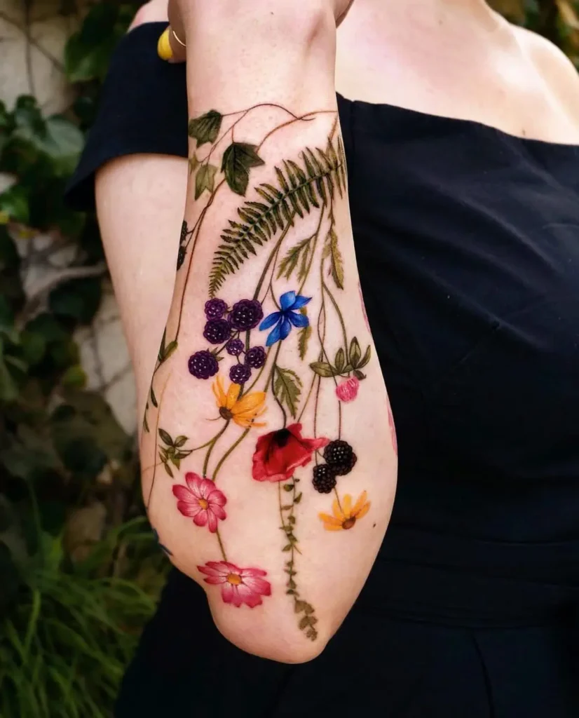 Colorful sleeve tattoo with wildflowers, blackberries, ferns, ivy, and trailing vines on a woman’s forearm.