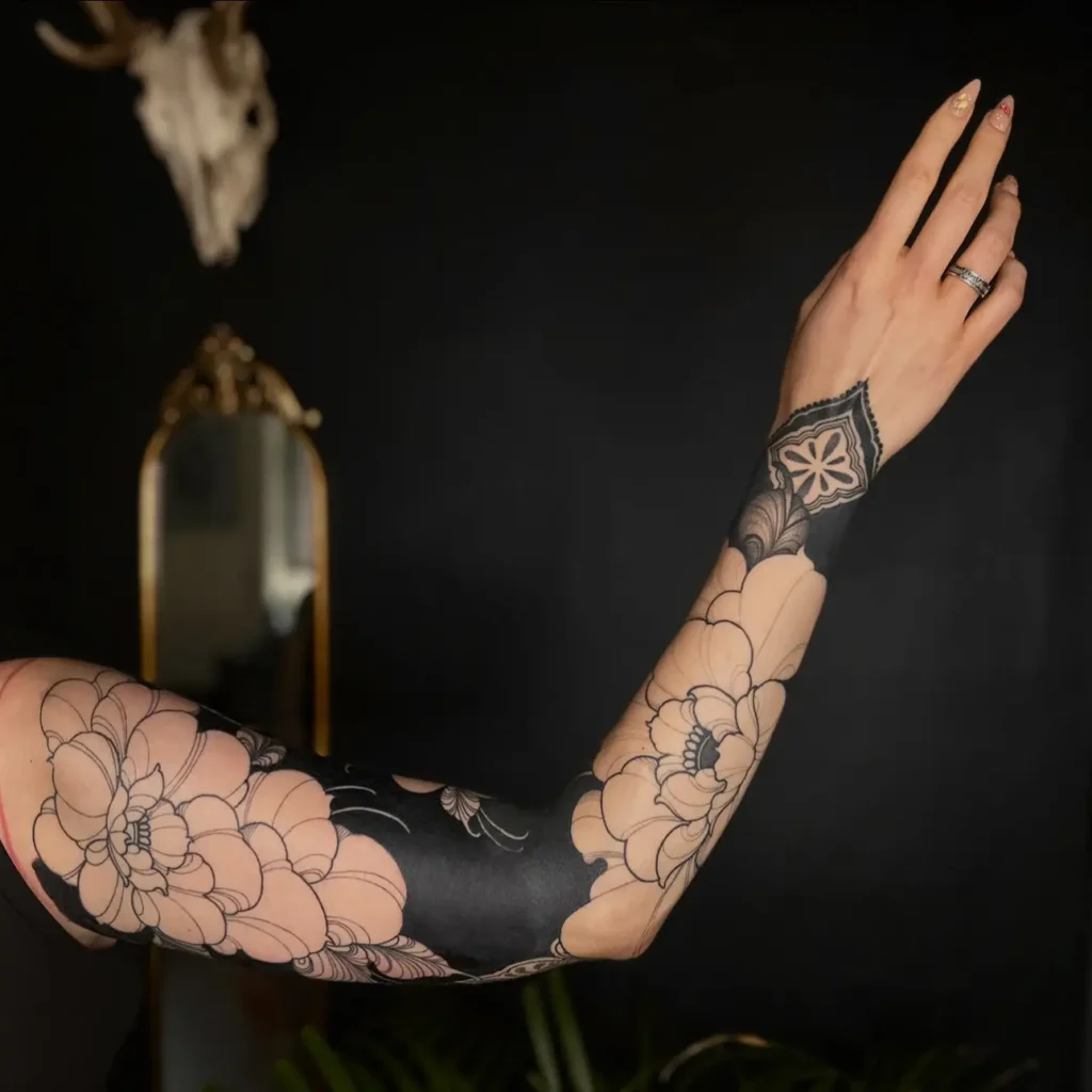 Blackwork sleeve tattoo with outlined peonies and a lace-inspired wrist design on a woman’s arm.