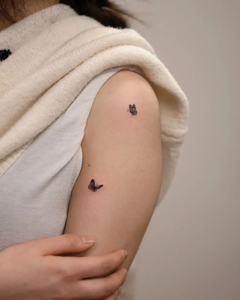 A minimalist tattoo with two small black butterflies on the upper arm, symbolizing transformation, freedom, and hope.