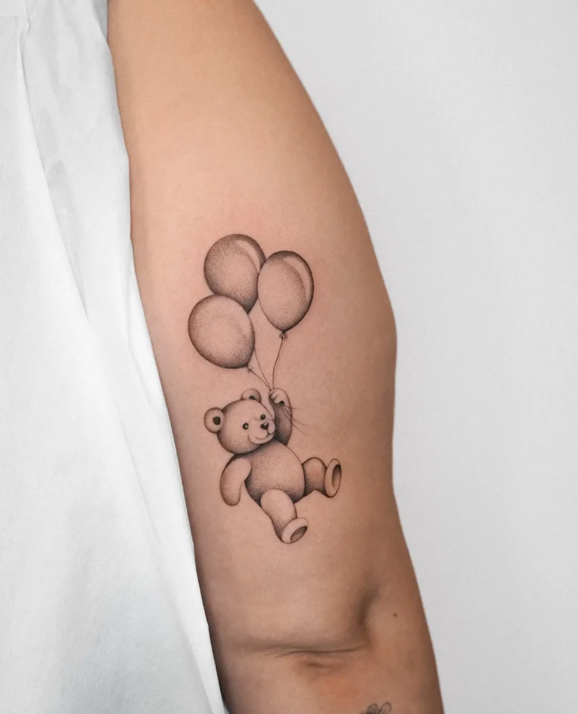 Upper arm tattoo for women featuring a cute teddy bear holding three balloons—softly shaded, playful, and nostalgic design.
