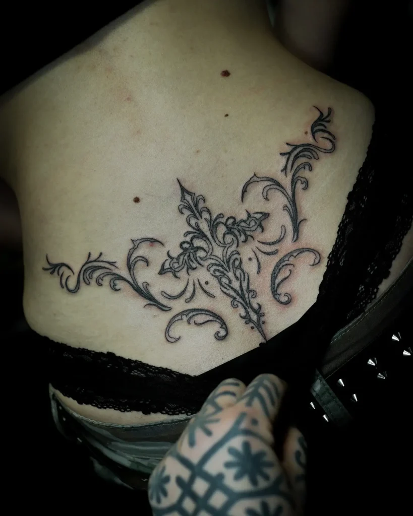  Lower back tattoo with intricate scrollwork and filigree, designed in a vintage, ornamental style with fine black lines.