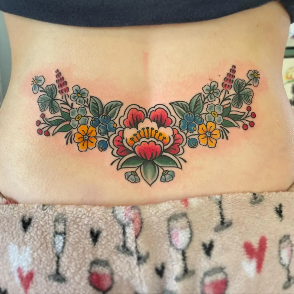 Lower back tattoo with bright folk-art flowers and leaves in red, yellow, blue, and green, outlined in bold black ink.