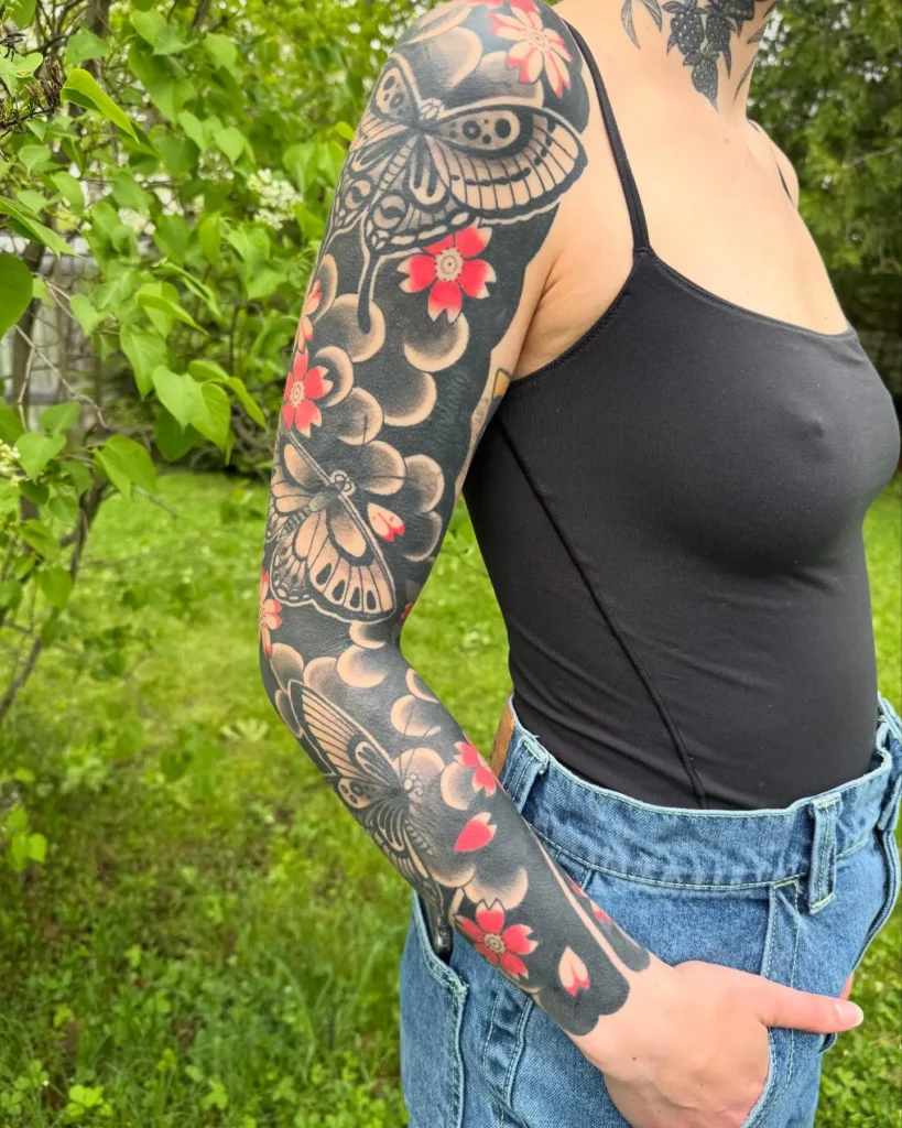 Japanese-style sleeve tattoo with blackwork butterflies, red cherry blossoms, and gray swirling clouds on a woman’s arm.