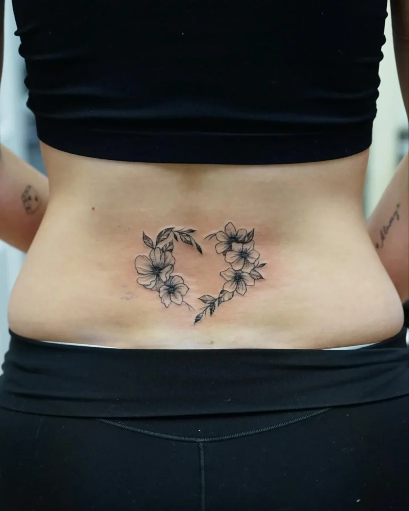 Lower back tattoo with a heart shape formed by delicate flowers and leaves, created in fine black linework.