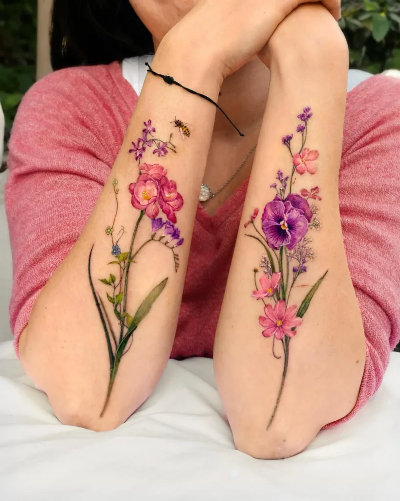 Colorful floral sleeve tattoos on both forearms featuring pink, violet, and purple flowers with greenery and a small bee in watercolor style.