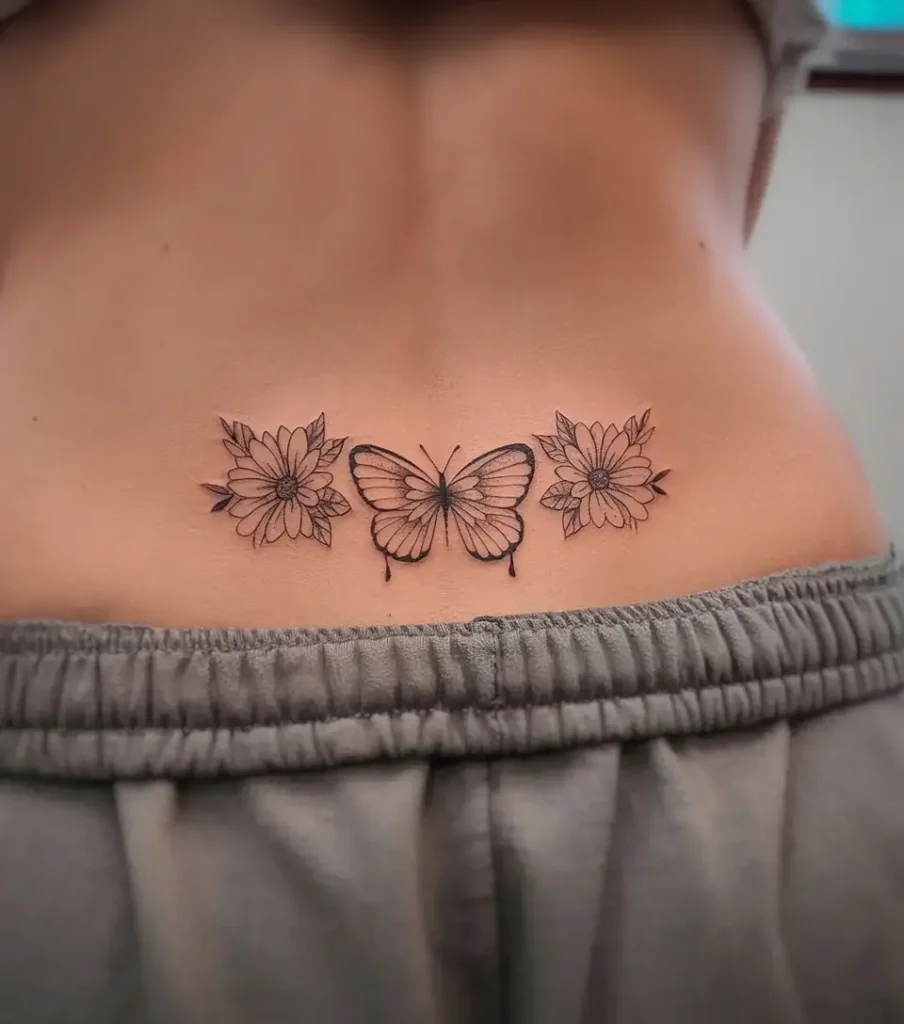 Lower back tattoo with a detailed butterfly centered between two blooming flowers, designed with fine black linework.
