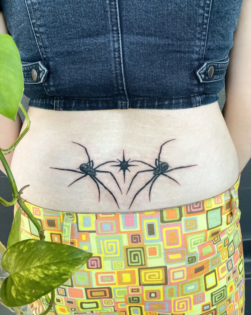 Lower back tattoo with two black spiders and a central starburst, drawn in fine black lines with a symmetrical layout.