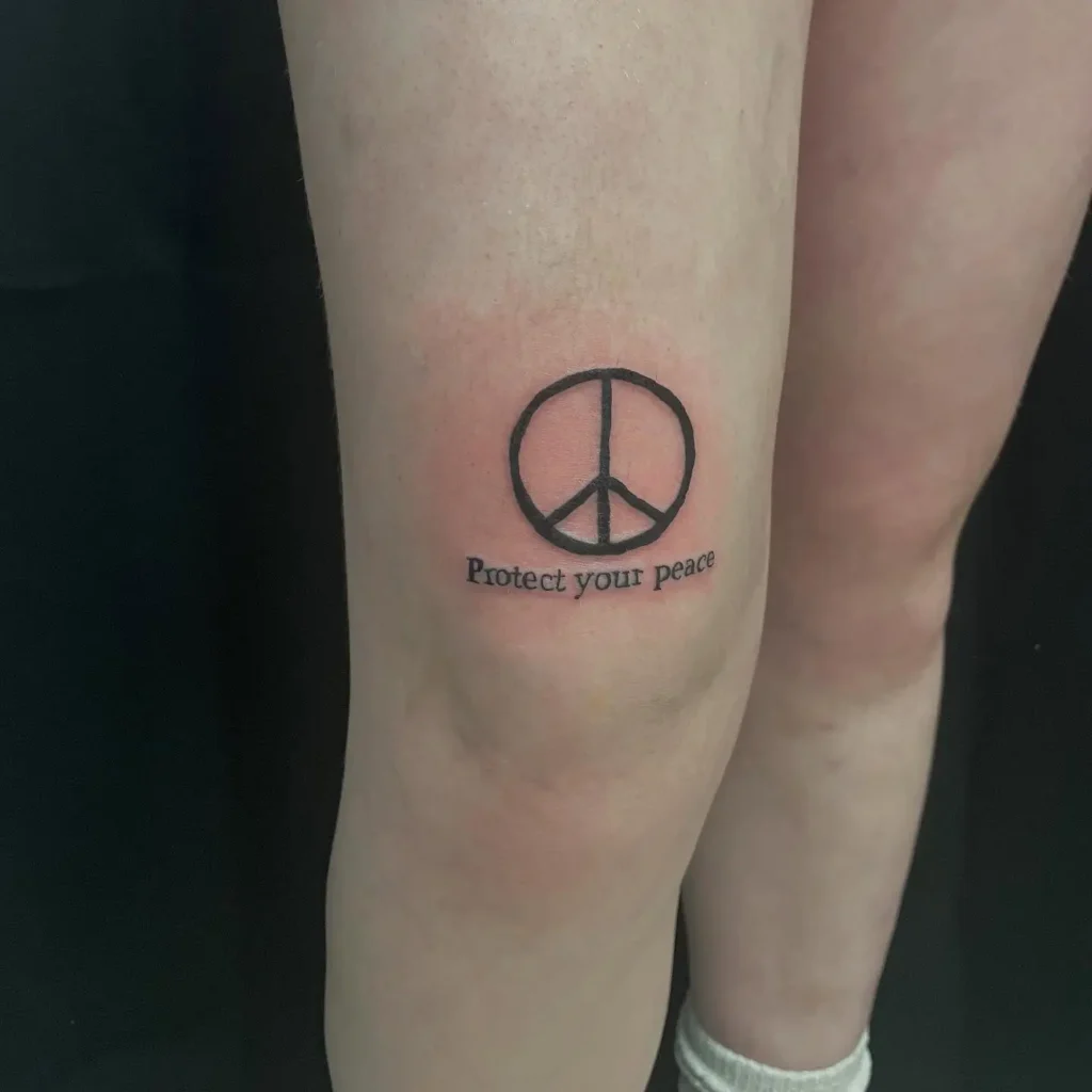 A tattoo above the knee featuring a black peace symbol with the words “Protect your peace” written underneath, representing mental health and self-care.