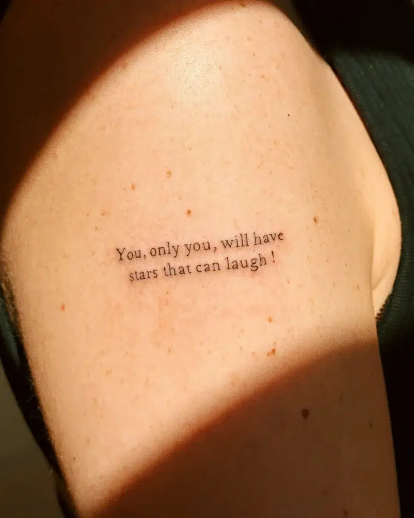 A tattoo on the upper arm with the phrase “You, only you, will have stars that can laugh!” in fine black script, representing hope and positivity.