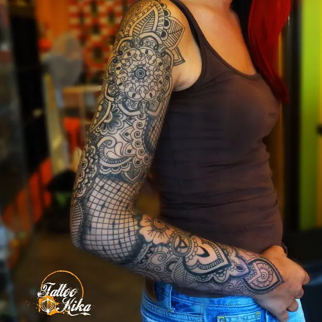  Full sleeve tattoo with black ink mandalas, floral lacework, and geometric grid patterns covering a woman’s entire arm.