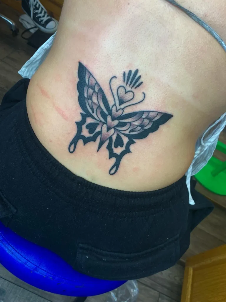 Lower back tattoo of a butterfly with bold black lines and heart motifs within its wings.