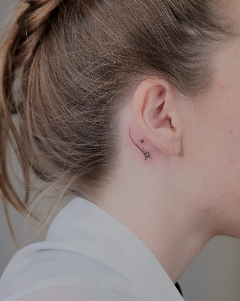 A fine line tattoo of two small shooting stars placed behind a woman's ear. The design features a soft curved line ending in minimal starbursts, complementing her tied-back hair and fair skin tone.








