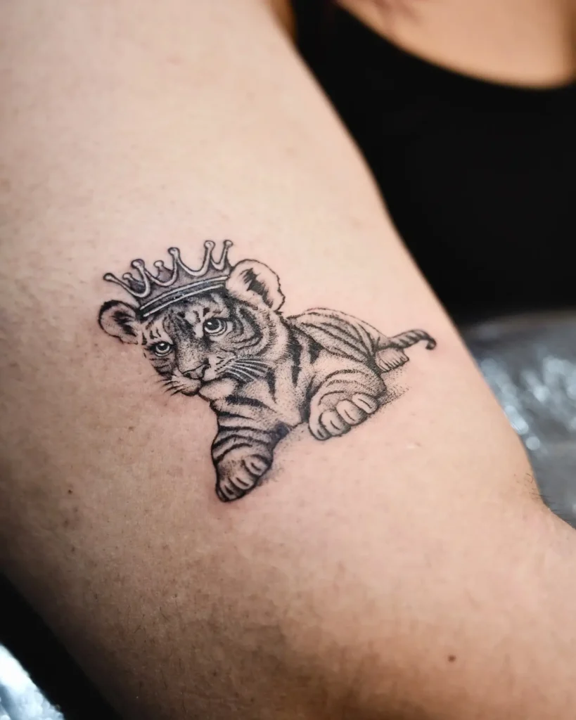 Small tattoo of a baby tiger cub wearing a royal crown, inked in black and gray fine lines on upper arm. Features soft shading and expressive details, capturing the cub’s gentle yet noble presence. A cute and meaningful choice among animal tattoo ideas simple, symbolizing strength, potential, and royalty. Suitable for small arm or shoulder tattoos.








