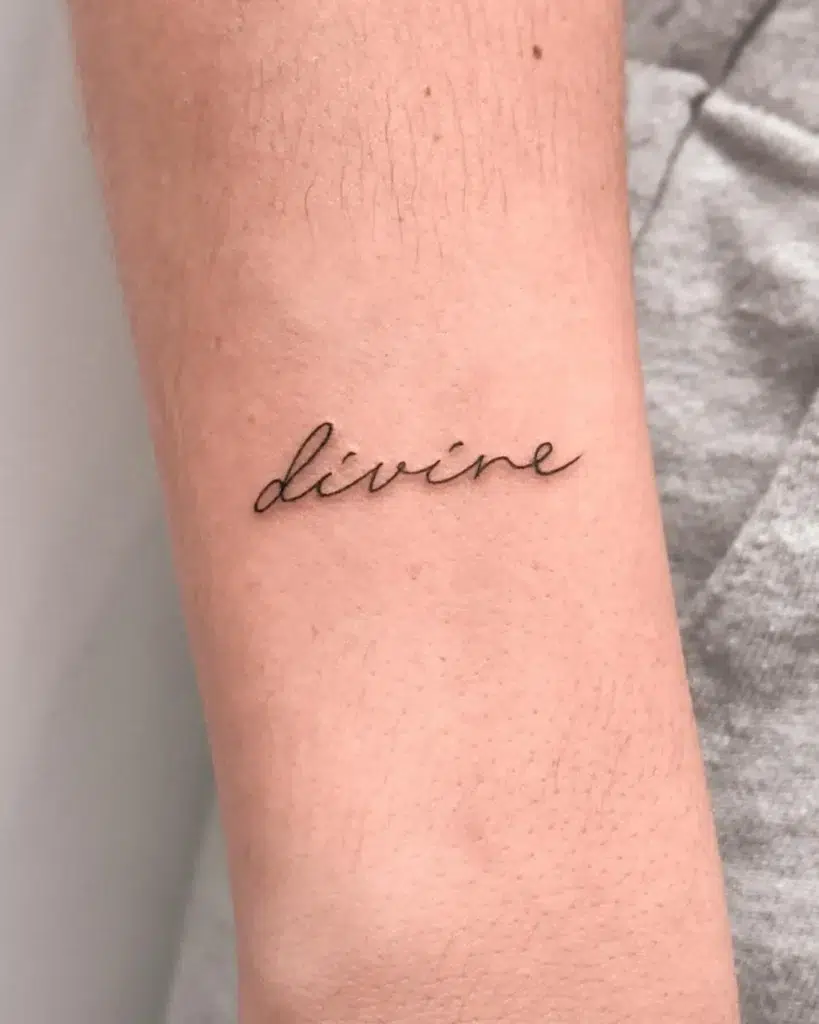 Minimalist cursive fine line tattoo of the word “divine” on the forearm, symbolizing grace, self-worth, and inner strength.