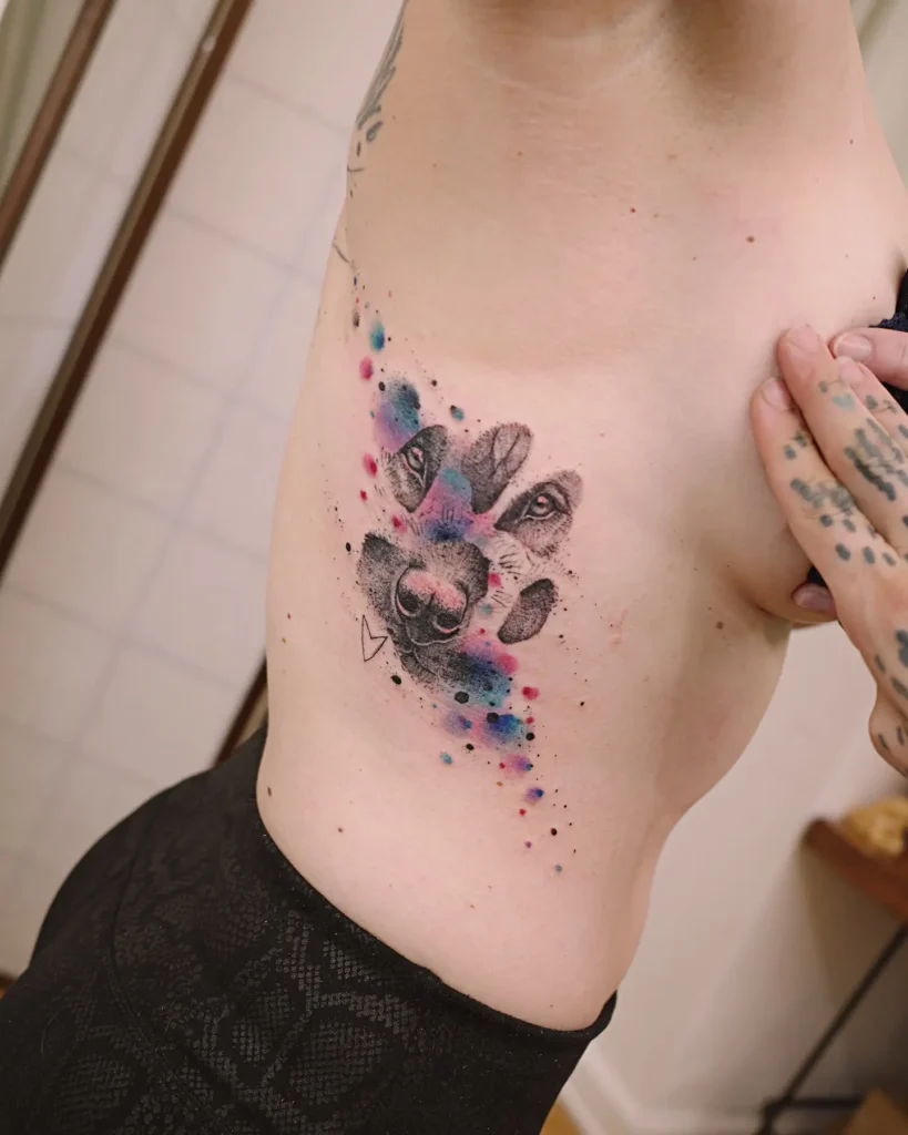 Watercolor dog tattoo featuring a paw print with a dog’s face and colorful splashes of pink, blue, and purple, accented with fine dots and a small heart.