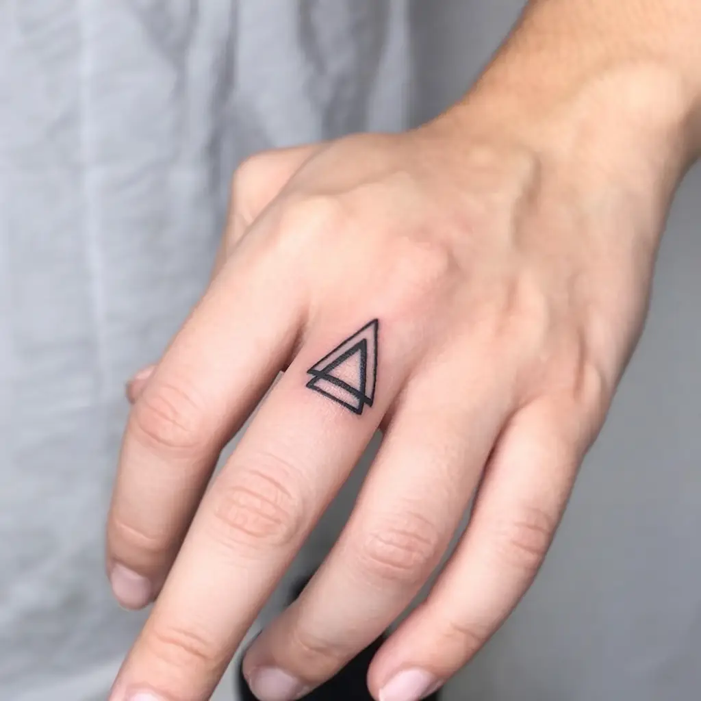Close-up of a hand with a minimalist geometric triangle tattoo on the ring finger. The black ink design features two overlapping outlined triangles, creating a bold and modern aesthetic. Clean lines and simple symmetry give the tattoo a sleek, contemporary look. Ideal for those who appreciate abstract art, symbolism, and small finger tattoos with a subtle yet edgy vibe.









