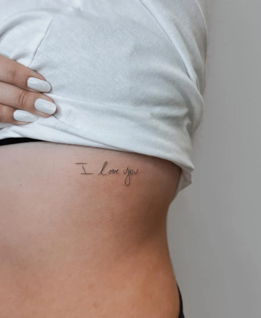 Fine line handwritten “I love you” tattoo on the ribcage, symbolizing emotional connection, love, and intimacy.