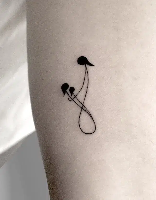 Minimalist black ink tattoo featuring an abstract infinity symbol created with a single continuous line, forming three stylized figures to represent a parent and two children. The design’s simplicity highlights eternal family connection and love in a subtle, modern style.