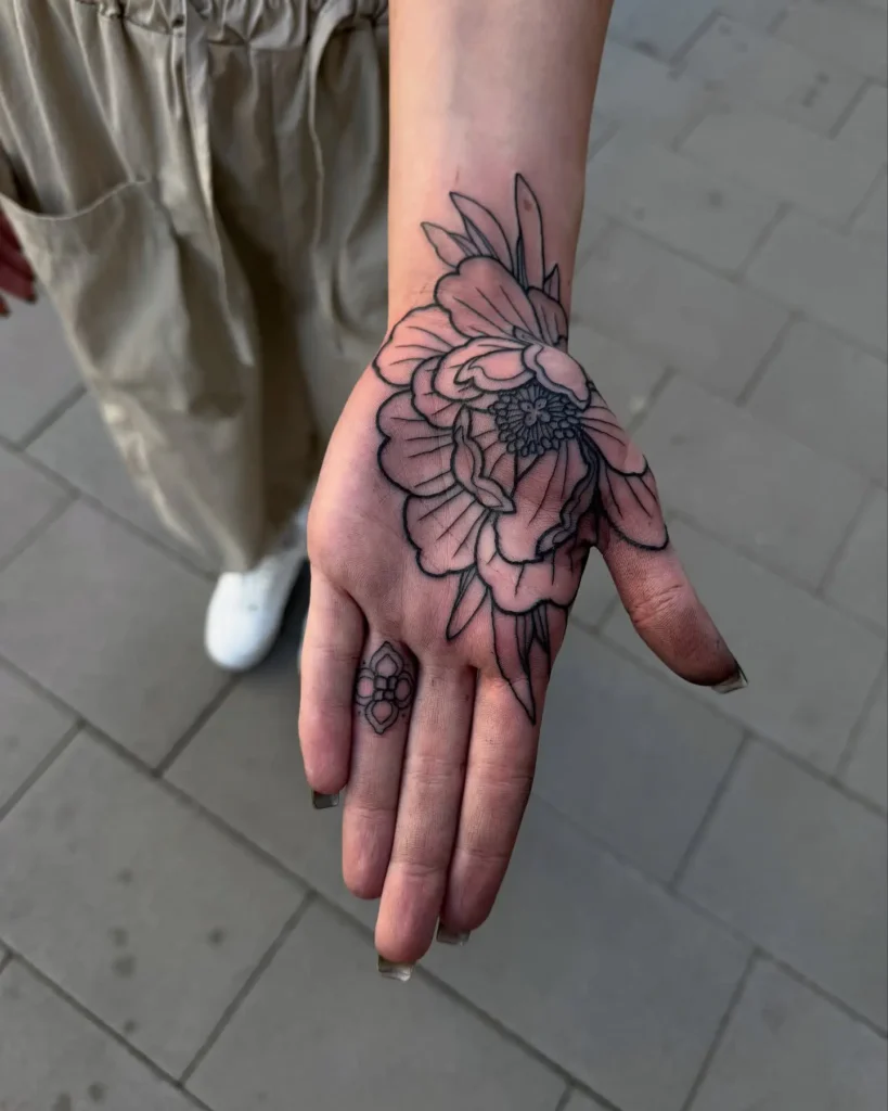 A hand with a peony blossom tattoo, featuring detailed petals and finger geometric shapes, set against a tiled background.