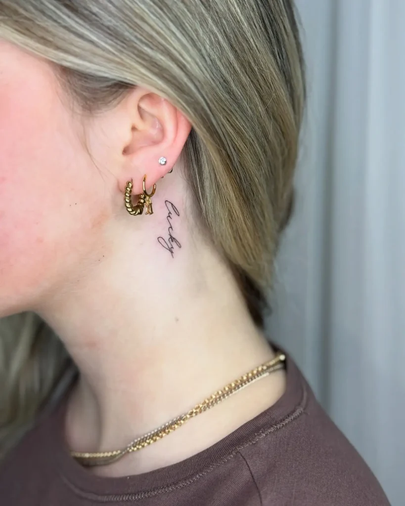 A minimalist behind the ear tattoo of the word "lucky" in fine cursive script on a woman’s neck, styled with blonde hair, gold hoop earrings, and a layered gold necklace.