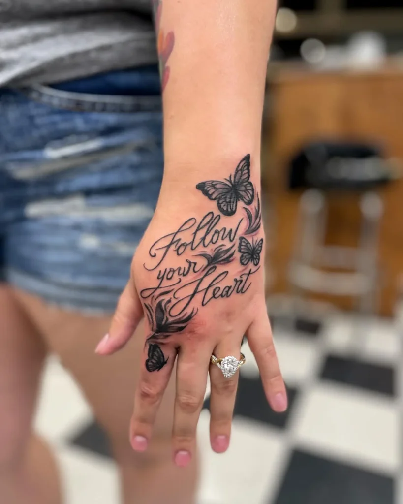 A hand with a "Follow your heart" tattoo in elegant script, featuring butterflies and vines, paired with a sparkling ring against a checkered background.