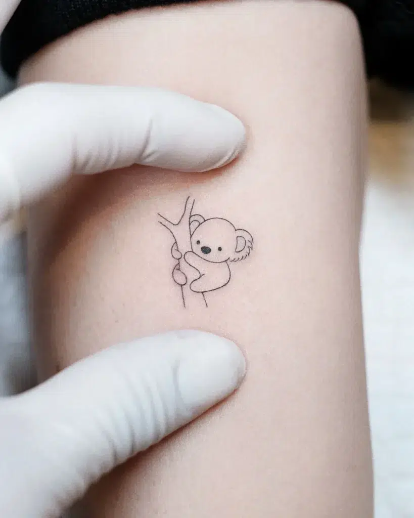 Tiny koala line tattoo on arm showing a cute koala peeking from behind a tree branch. Minimalist design with soft curves and clean lines, creating a sweet and subtle animal tattoo idea perfect for delicate placements or micro tattoos
