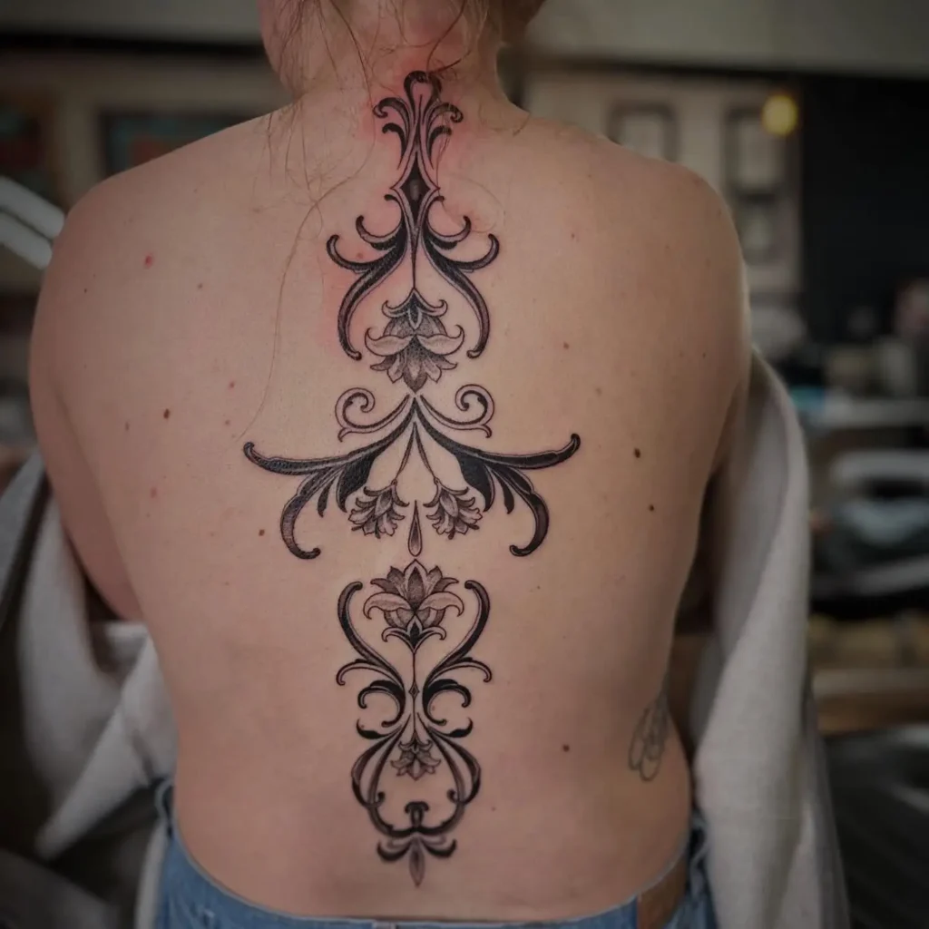A large, ornate baroque spine tattoo with bold swirling motifs and floral accents running down a woman’s back.