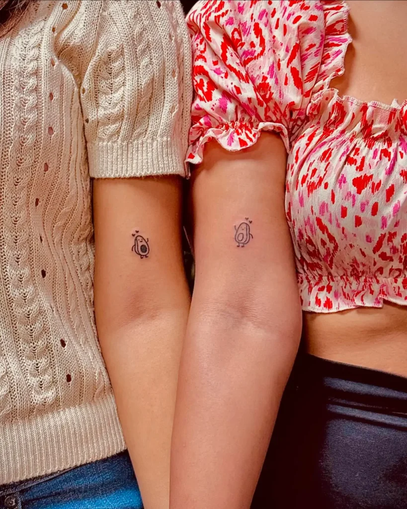 Matching tattoos on two arms, each showing a small, simple avocado character with a tiny heart above. The avocados are drawn in black ink with rounded shapes, adding a cute and whimsical touch. This unique design captures the joyful spirit of mom and daughter tattoos, focusing on love, connection, and fun memories.