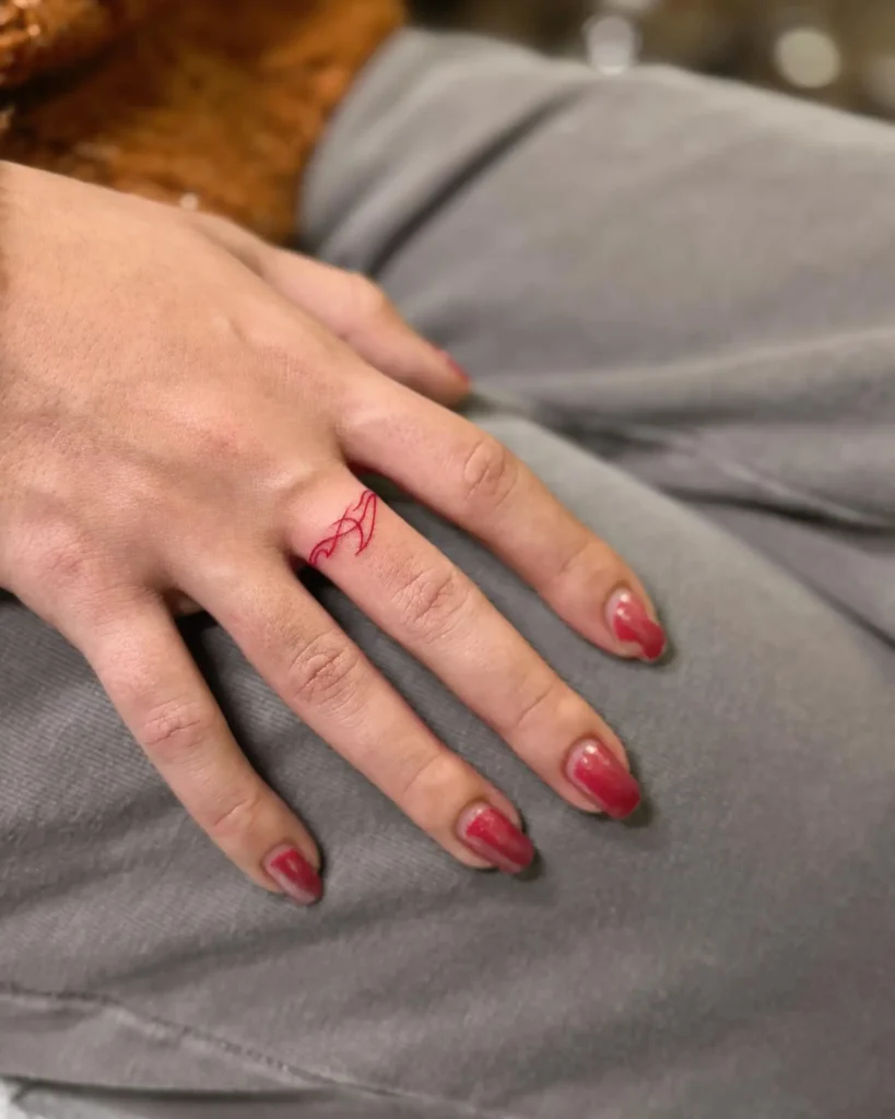 Close-up of a woman's hand showcasing a unique red ink finger tattoo with an abstract line art design on the middle finger. The fine-line tattoo wraps gently around the finger, creating a modern and stylish look. Her nails are painted with a glossy red polish, complementing the bold tattoo color. This design is perfect for women looking for minimalist and artistic finger tattoos with a pop of color.









