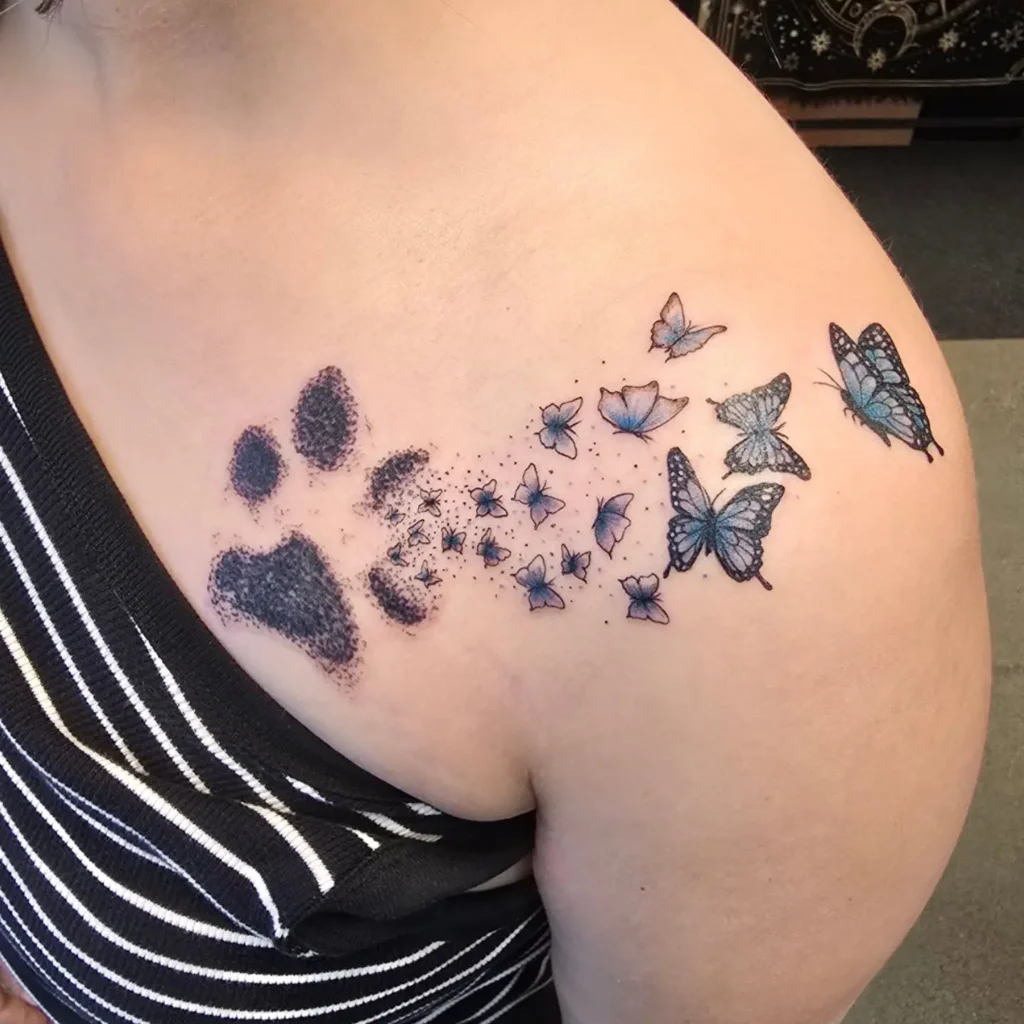 Tattoo of a black dog paw print fading into blue butterflies on the shoulder, symbolizing memorial and transformation in dog tattoo ideas.