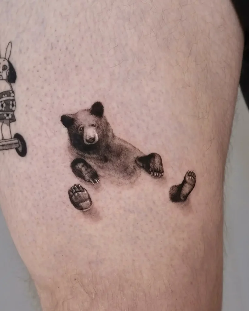 Tiny floating bear tattoo in black and grey style, with separate soft-shaded paws and head arranged as if suspended midair. Minimalist and surreal placement on upper thigh, blending realism with artistic abstraction. Playful tone with light detailing.