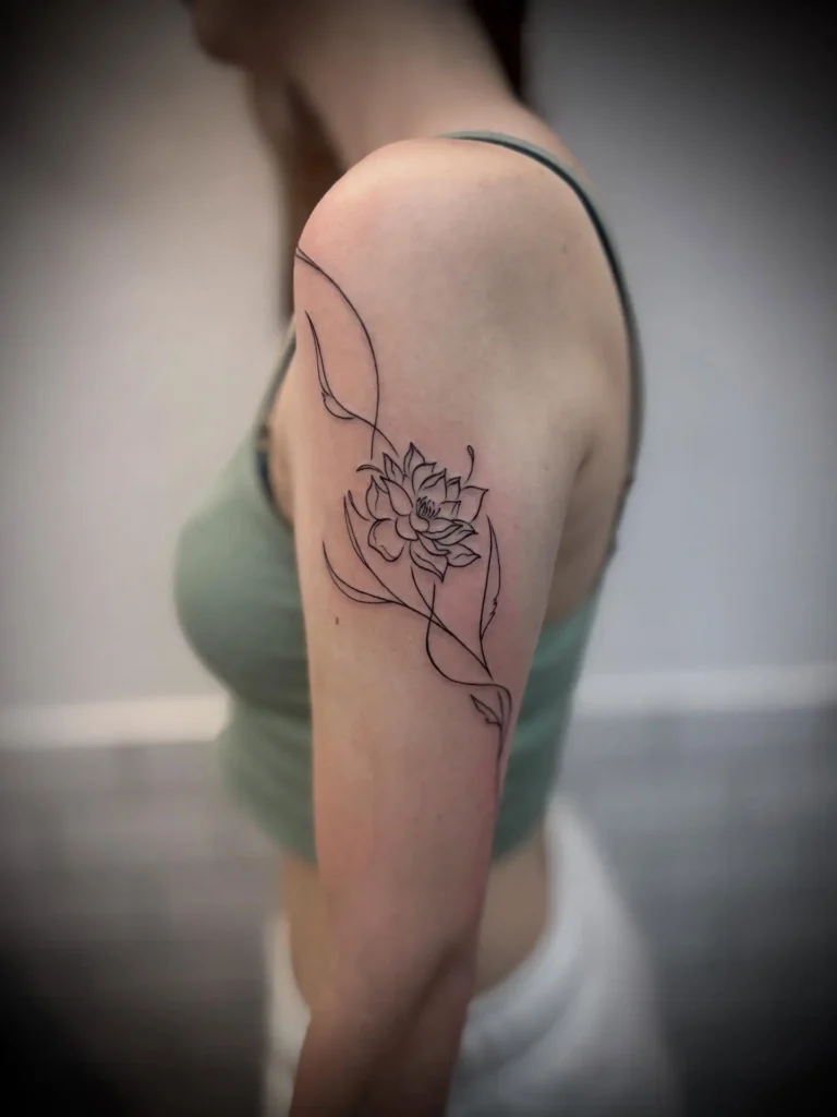 Black line art tattoo of a lotus flower with leaves, wrapping around a woman’s upper arm.