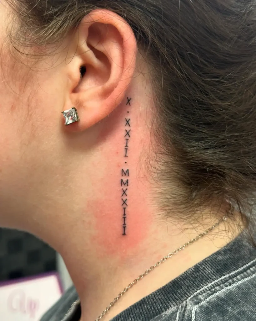 A clean black Roman numeral tattoo placed vertically behind a woman’s ear, spaced with small dots, symbolizing a significant date. The skin around the ink appears slightly red and freshly done.









