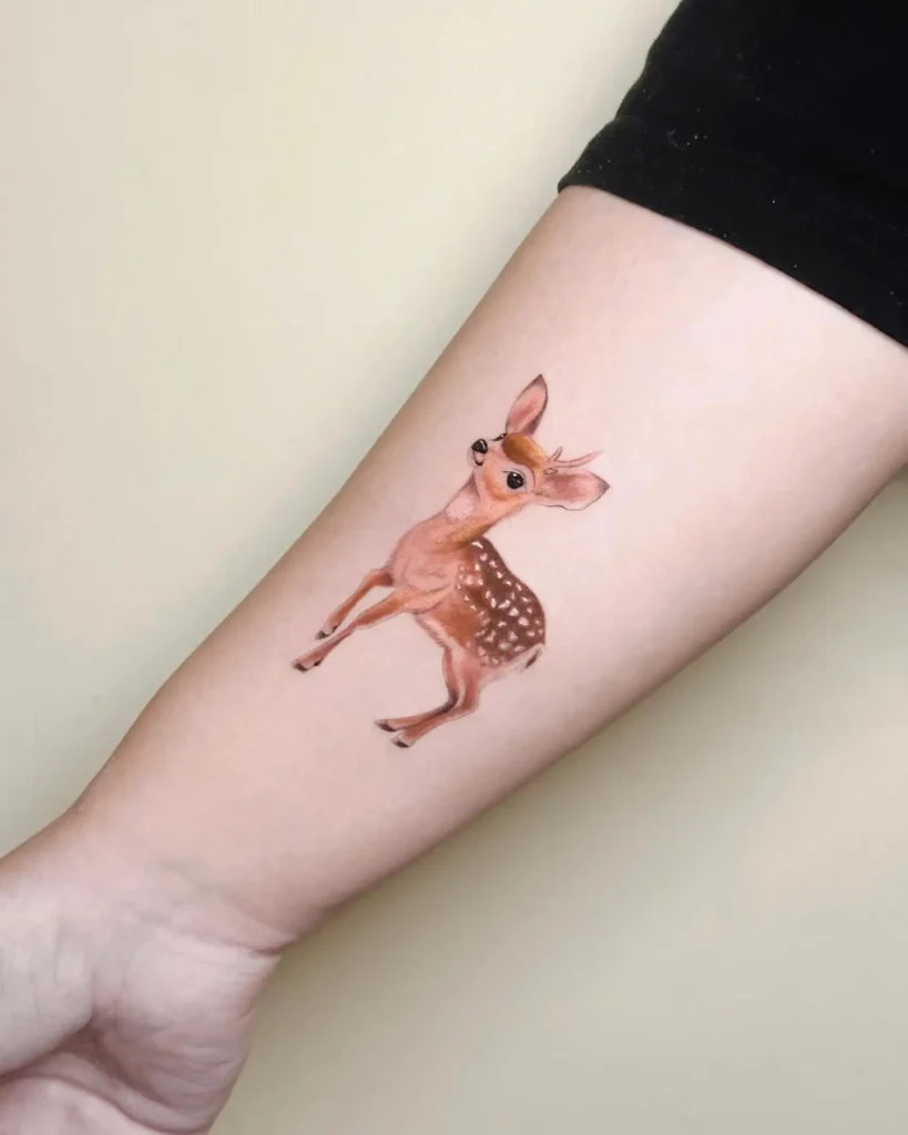 Realistic tattoo of a young fawn placed on the inner forearm, featuring soft brown shading, white spots, and a wide-eyed, curious look. The design captures a gentle, storybook-like innocence with smooth, detailed coloring and placement that enhances the natural curve of the arm.