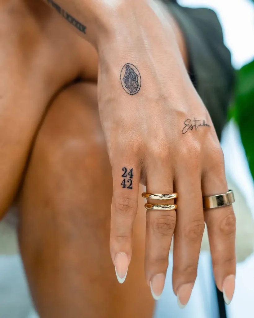 A hand with a minimalist Virgin Mary tattoo, "24:42" finger number, and "Esther" script, paired with gold rings against a soft background.