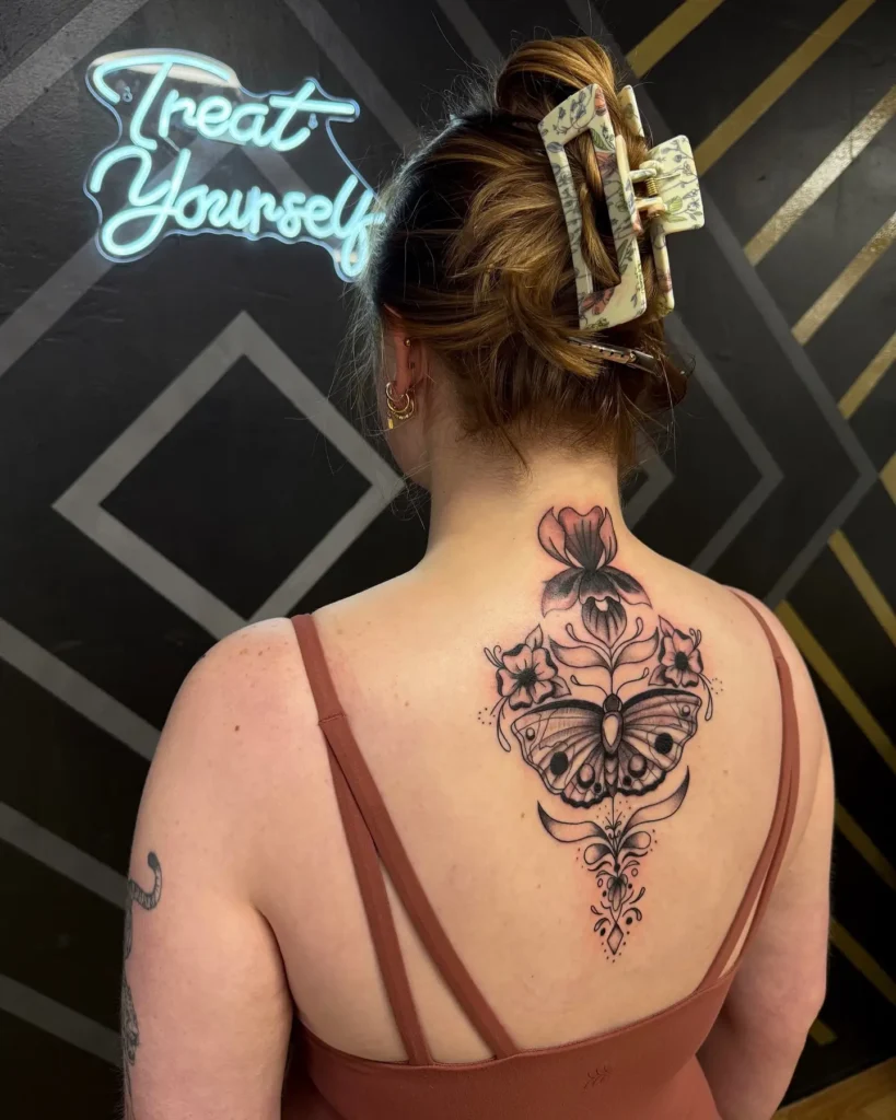 A spine tattoo with a detailed moth and blossoming flowers, featuring fine lines and shading, on a woman’s upper back.