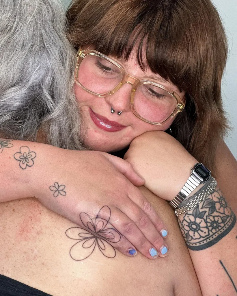 Close-up of a hand resting gently on someone’s shoulder, showing fresh and healed flower outline tattoos on the hand and wrist. The overlapping petals create a soft, organic pattern in black ink. This tattoo style stands out for its simplicity and meaning, making it a sweet option for mom and daughter tattoos that celebrate togetherness and cherished memories.