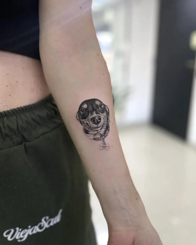 Realistic tattoo of a smiling dog’s face with a bone-shaped name tag, placed on the forearm, showcasing lifelike detail for personalized dog tattoo ideas.