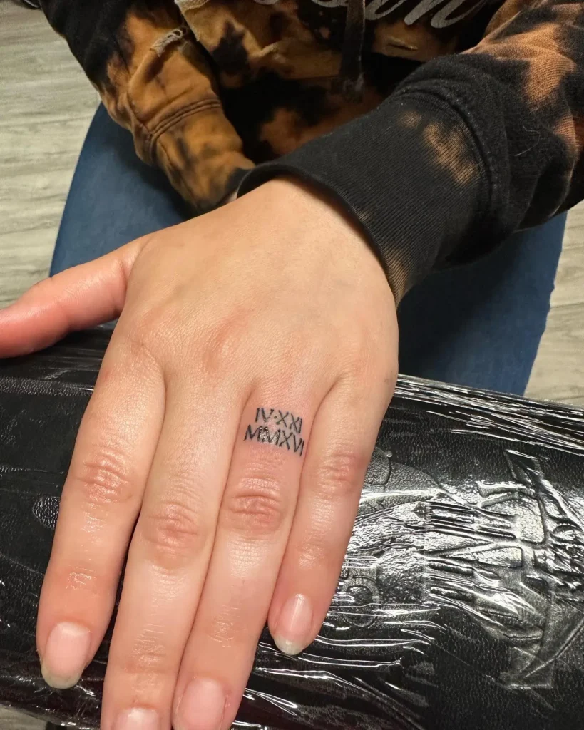 Close-up of a woman’s hand featuring a clean and bold Roman numeral finger tattoo on the ring finger. The black ink design reads “IV·XXI MMXXVI,” representing a meaningful date in a stylish and timeless format. The minimalist lines and centered placement give the tattoo a modern yet classic appeal. A great example of finger tattoos for women who want to honor a special date with subtle and personal symbolism.









