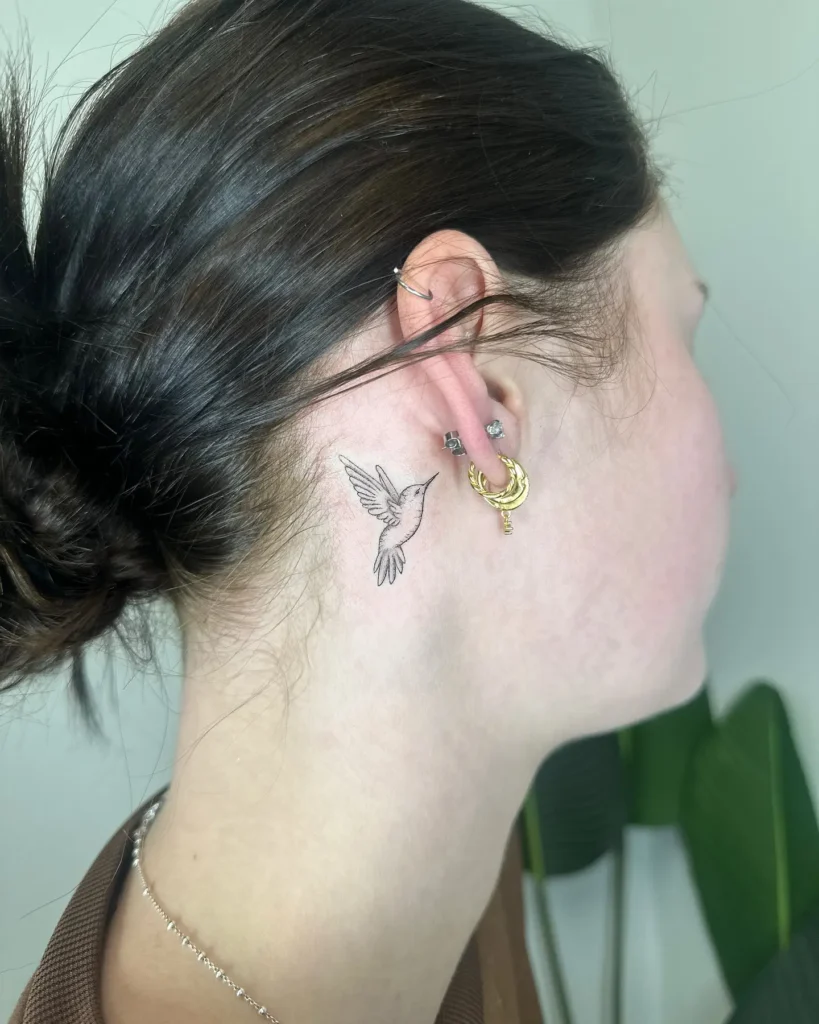 A behind the ear tattoo of a small, finely detailed hummingbird inked in black on a woman’s neck, paired with multiple gold and silver earrings and a soft bun hairstyle.









